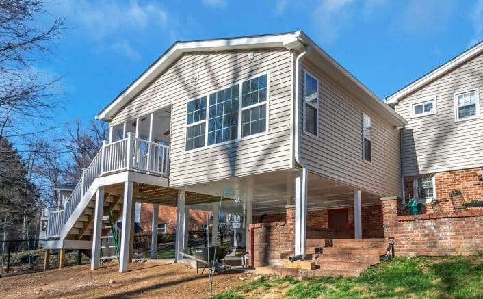 Rear view of a two-story house showcasing a new room addition on the first level, with a spacious deck above it, set against a backdrop of a well-kept lawn and clear blue sky