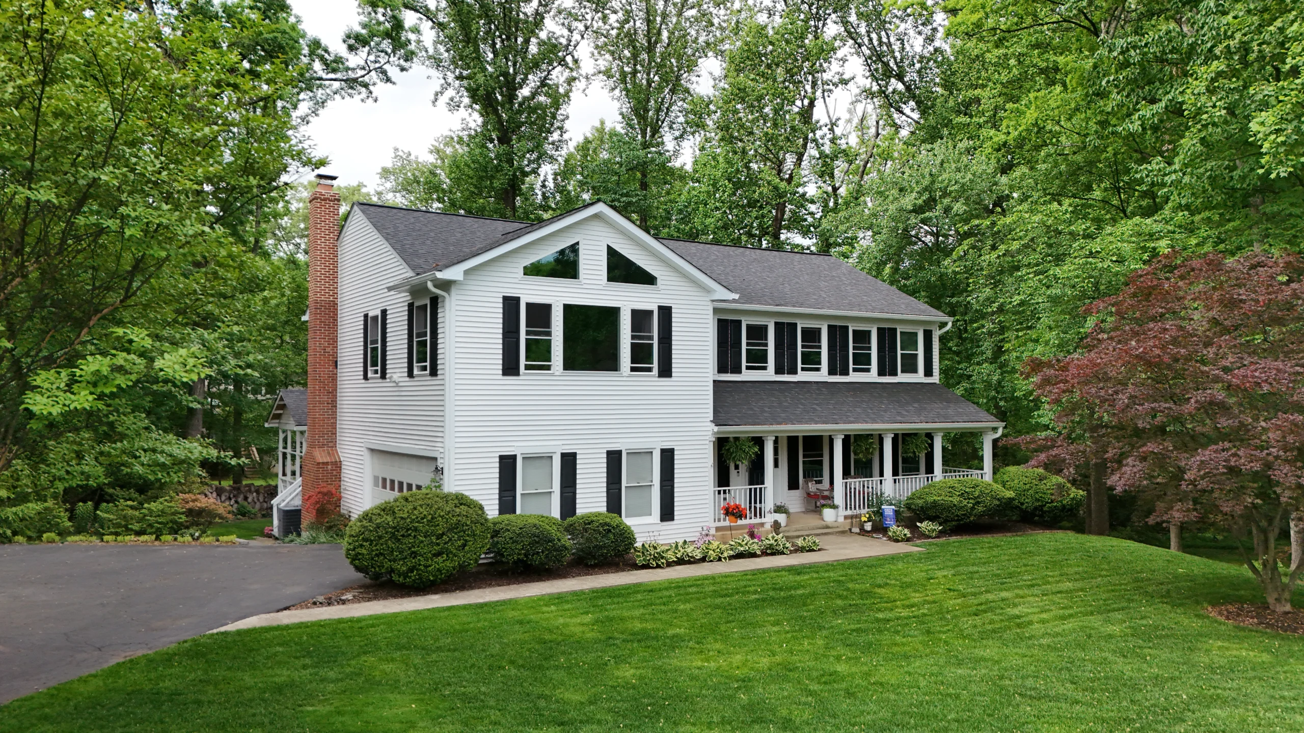 White two-story house with black shutters, brick chimney, and a covered porch amid a lush green yard and trees nearby.