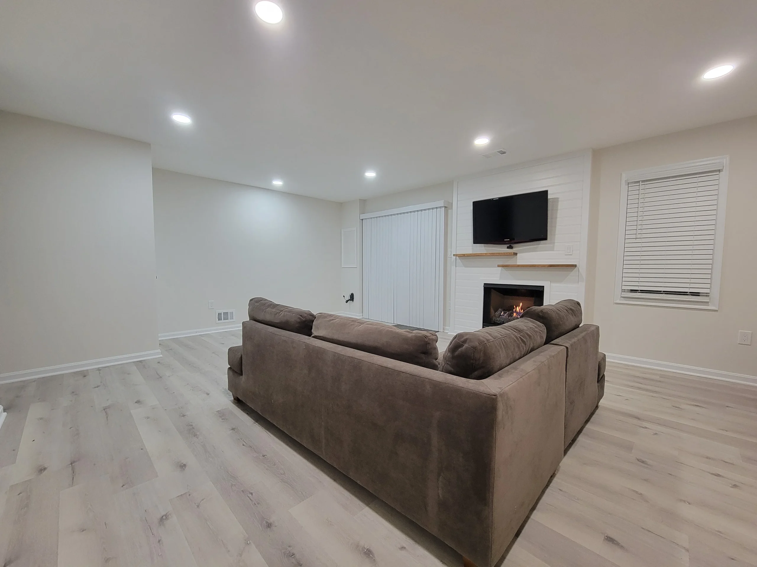 Cozy living room with a gray sectional sofa facing a fireplace and mounted TV on a white paneled wall, bright recessed ceiling lights.