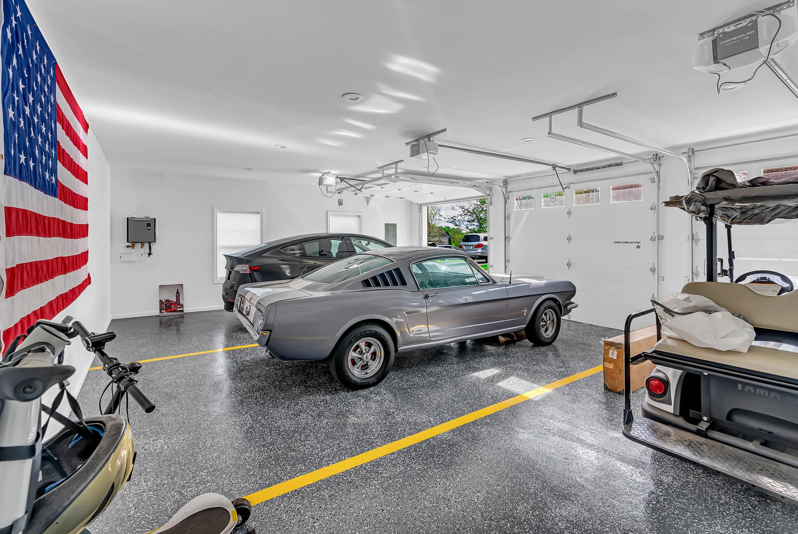 Garage with a gray vintage Mustang in the foreground, American flag on the left wall, and another car in the background inside a clean, organized space.