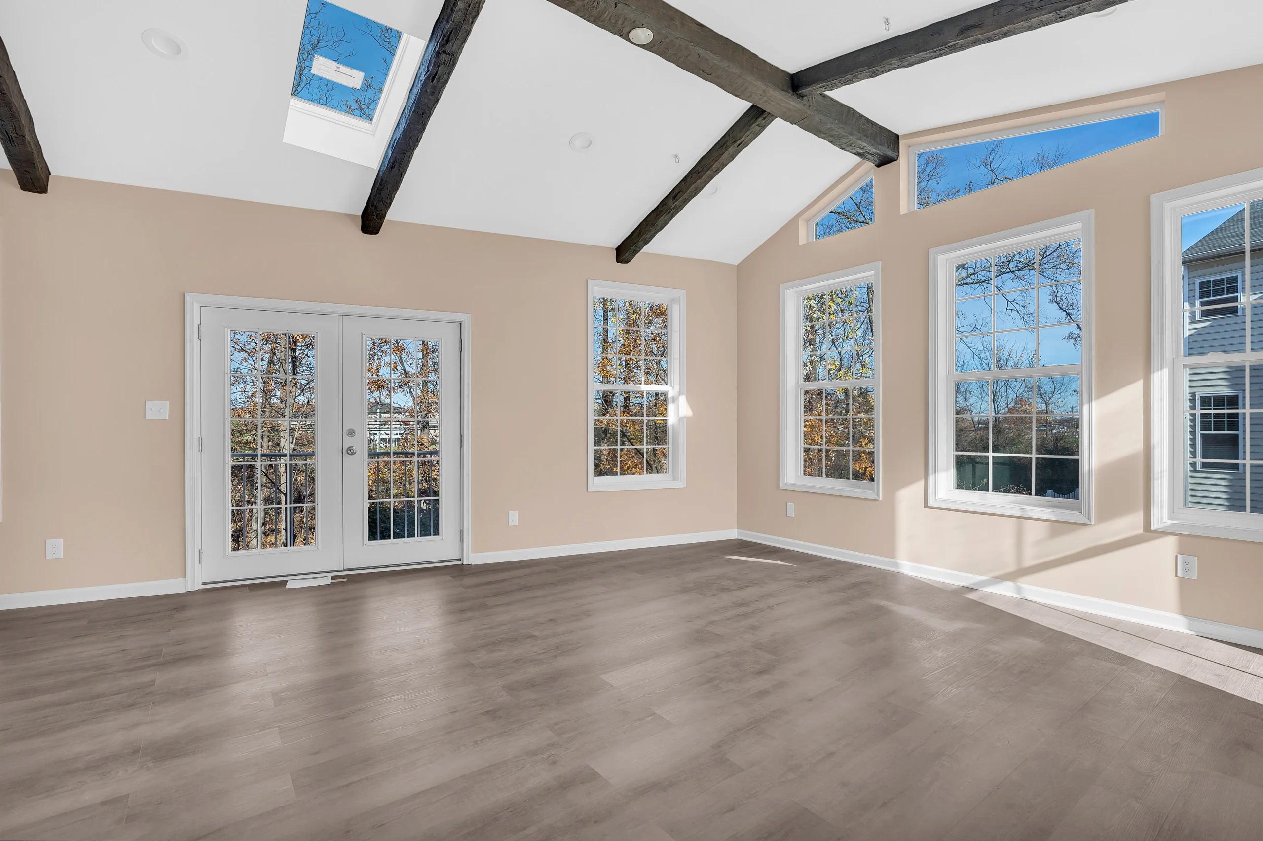 fairfax-station-extension-design - Mosaic Build - Mosaic Build Bright empty living room with beige walls, dark exposed ceiling beams, and large windows letting in sunlight; glass doors to outside.