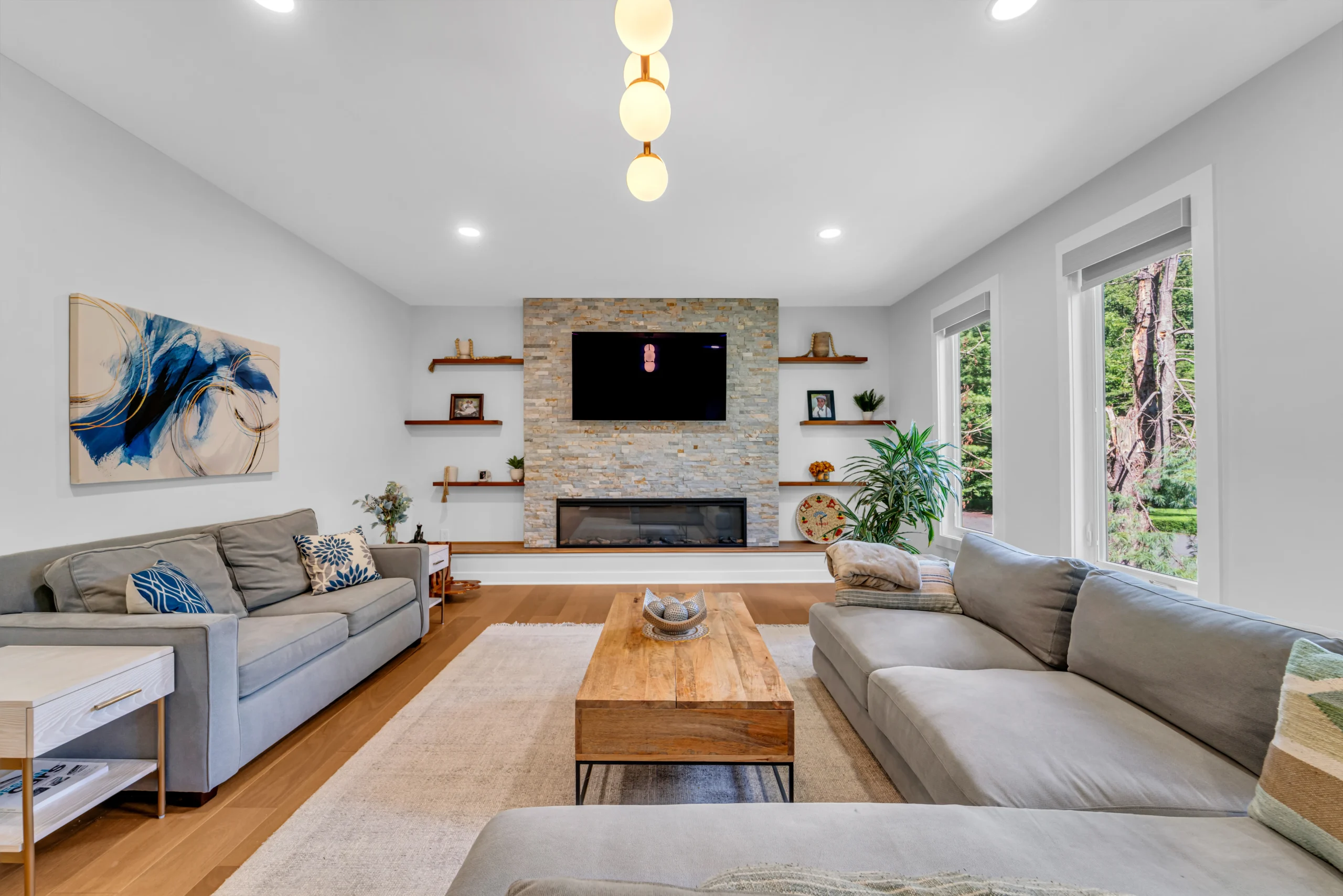 fairfax-station-home-addition - Mosaic Build - Mosaic Build Bright living room with a gray sectional, stone fireplace, and large windows showing greenery outside.