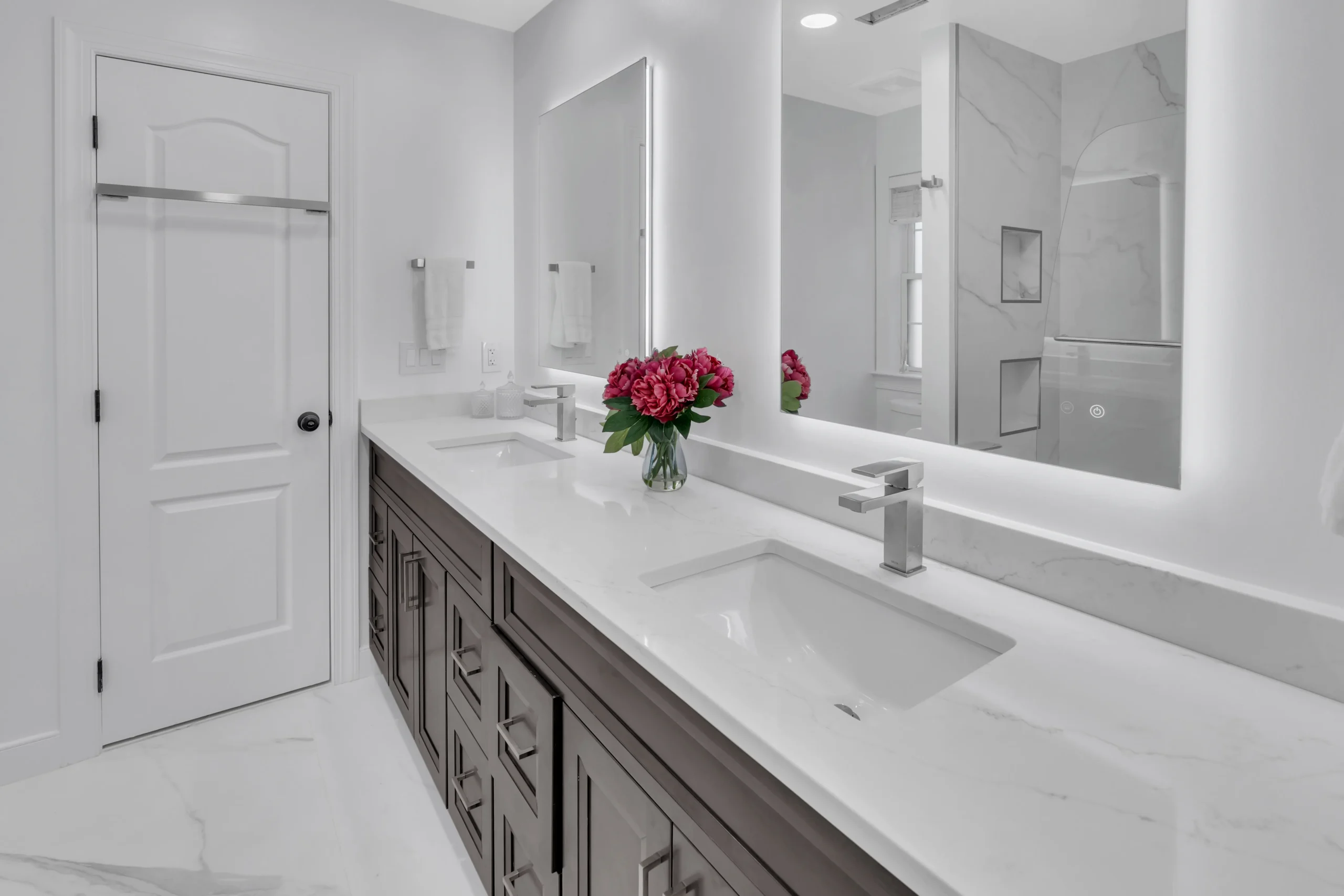 Modern white bathroom with double-sink vanity, dark wood cabinets, backlit mirror, and pink bouquet on marble countertop.